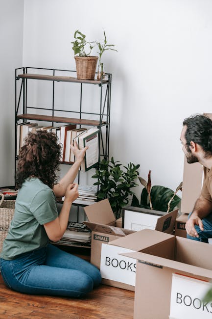 A man and a woman inside a room filled with packed cardboard boxes, some labeled with items such as 'books', during a home relocation. The man, wearing a grey hoodie, is sitting on the floor with a sl