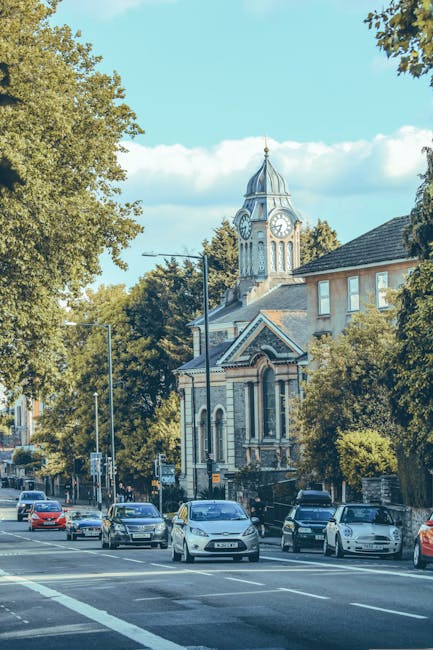 A view of a busy street in Putney with multiple parked and moving cars, including sedans and hatchbacks, along the roadside pavement. Behind the vehicles, there is a historic church with a prominent c